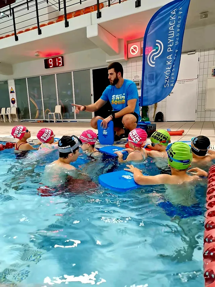 Instruktor w koszulce Swim Squad prowadzi zajęcia z dziećmi przy brzegu basenu